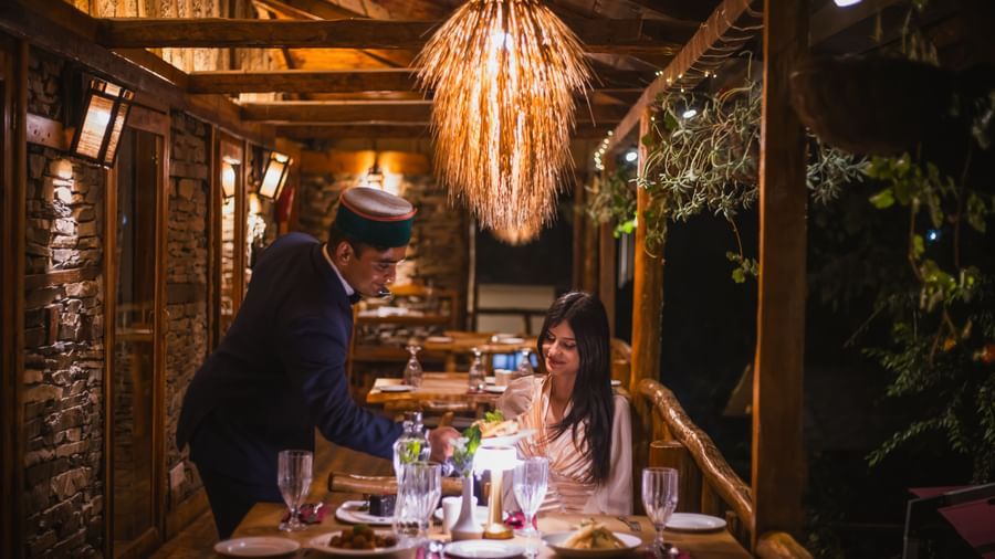 Outdoor dining experience under a glowing hanging lamp with guests enjoying a meal within a wooden pergola surrounded by greenery, highlighting the sophisticated and intimate culinary atmosphere at The Manor Sports & Wellness Hotel in Shimla.