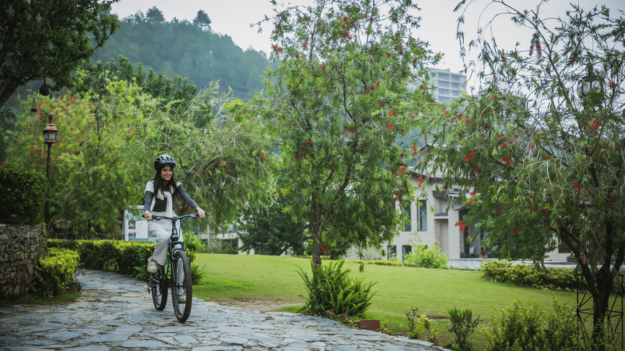 A woman riding a bicycle on stone pathway bordered by lush garden plants, with misty hills in the background, showcasing wellness and recreational activities in the scenic environment of The Manor Sports & Wellness Hotel in Shimla.