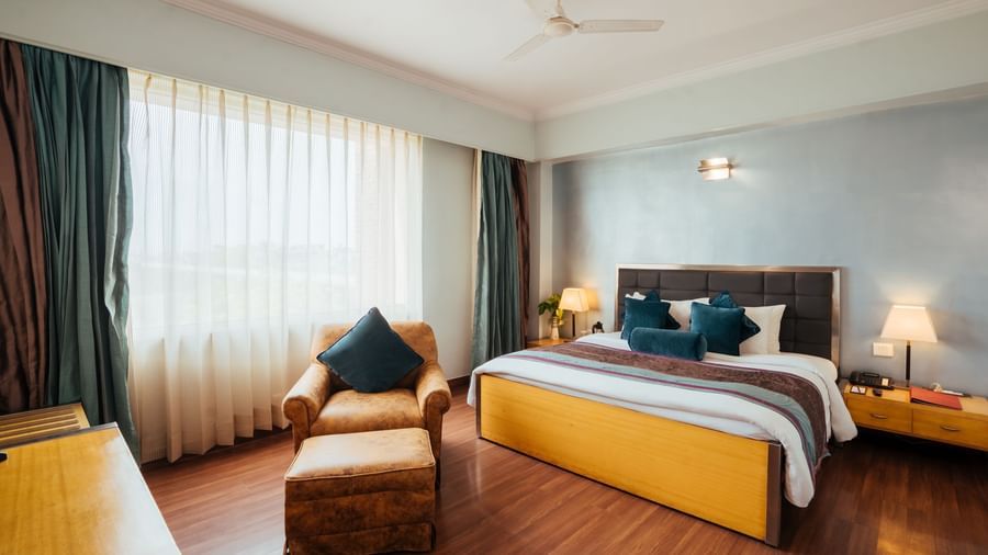 A room at The Piccadily Lucknow hotel featuring a double size bed, a sofa chair, bedside table with lamps on either side, and a large window with curtains.