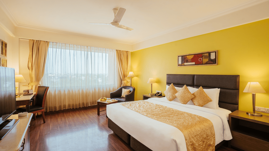 A spacious bedroom with a yellow feature wall, a bed with a golden runner, writing desk, armchair, and sheer window drapes at The Piccadilly Lucknow.