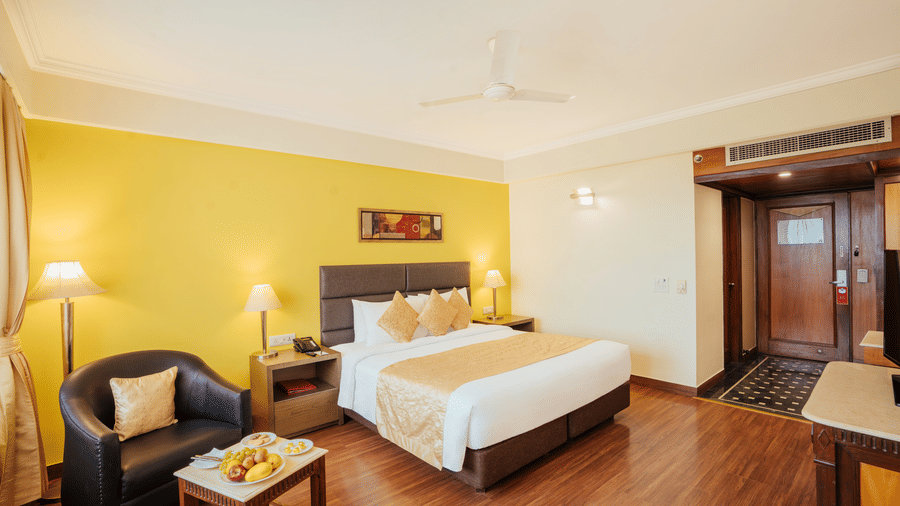 A modern bedroom with a neatly-made bed, yellow accent wall, leather armchair, coffee table with a fruit plate, and wooden flooring at The Piccadilly Lucknow.