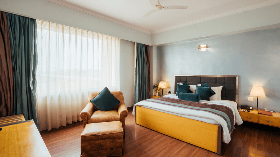 A bedroom at The Piccadily Lucknow featuring a bed, a sofa chair, bedside table with lamps on either side, and a large window with curtains.