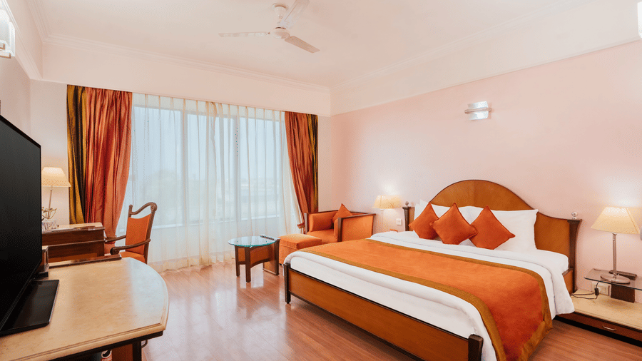 A warm-toned bedroom with a wooden bed, matching runner and cushions, work table, and a large window at The Piccadilly Lucknow.