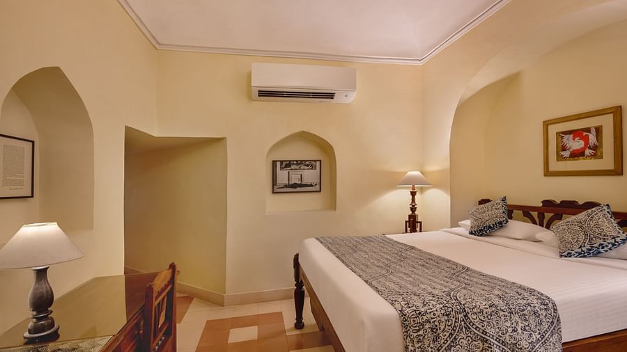 Room with bed, patterned runner and pillows, furniture including chair and lamps, arched alcoves, artwork, wall-mounted air conditioner, and patterned floor tiles - Tijara Fort-Palace - 19th Century, Alwar.