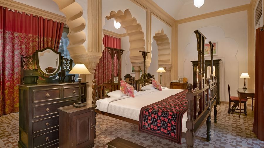 A room with a bed, a dresser with a mirror, a chair, a table with a lamp, and arched architectural details at Tijara Fort-Palace - 19th Century, Alwar.