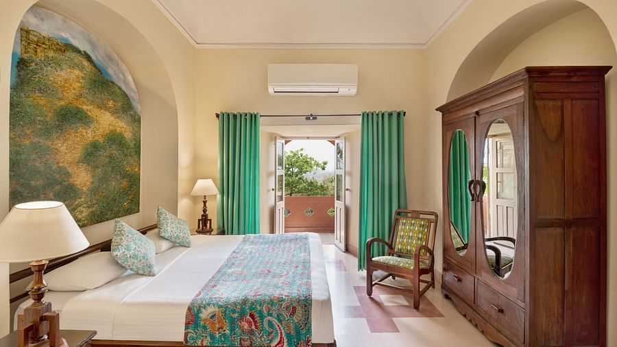 Bedroom with a bed, wooden wardrobe and chair, bedside tables with lamps, artwork, balcony with curtains, and AC at Tijara Fort-Palace, Alwar.
