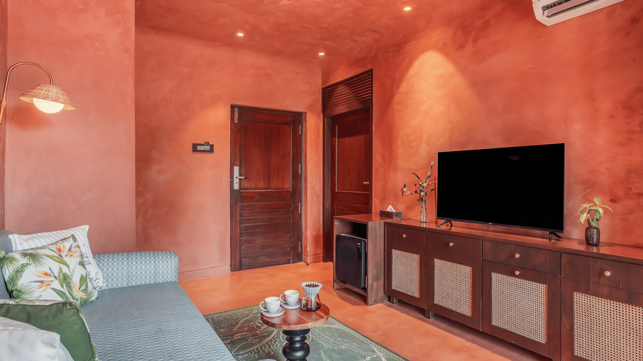 Living room at Unseen Boutique Hotel with reddish?orange walls, gray sofa, patterned cushions, wooden TV console, flat?screen television, and recessed ceiling lights.