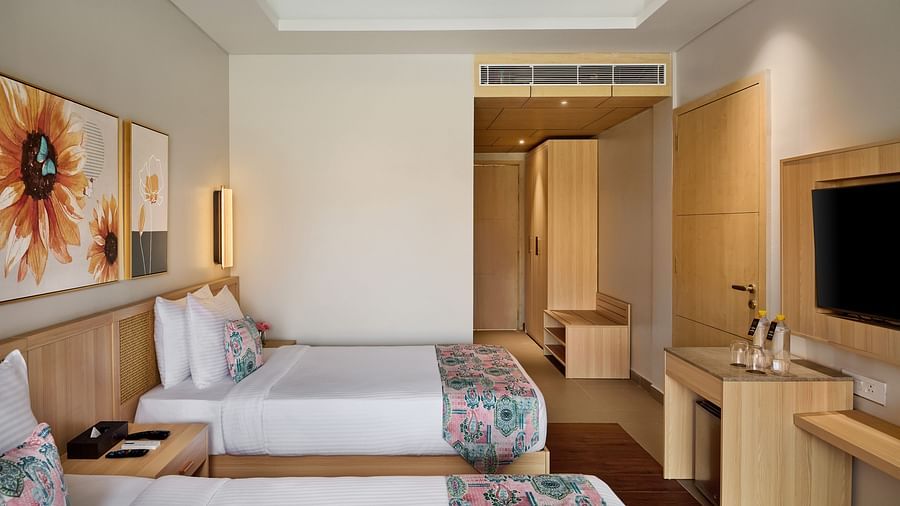 A hotel room features two beds with floral patterned bedspreads, flanked by a wooden desk and a nightstand. Sunflower paintings hang above each bed - Voyage The Solitaire Bliss.