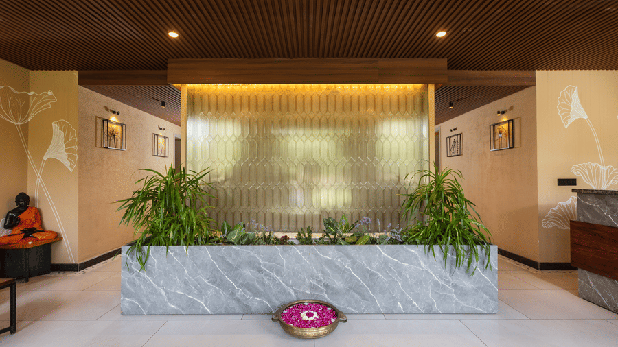 A modern spa lobby with a large,  water wall feature centered behind a white marble barrier a reception desk is visible in the background at The Golden Tusk, Jim Corbett
