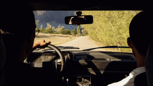 The view from inside a car shows a person driving on a road through a forested area.