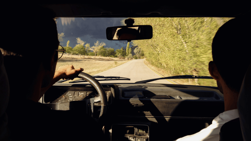 The view from inside a car shows a person driving on a road through a forested area.