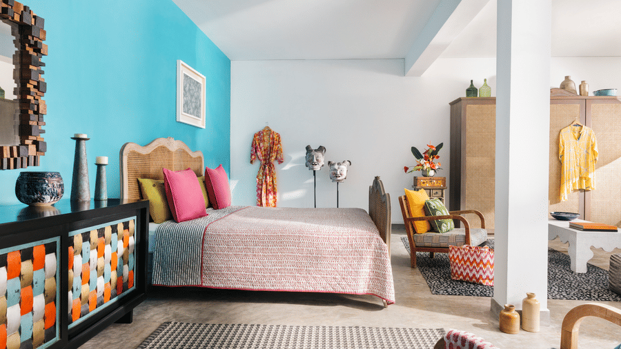 A spacious, brightly lit bedroom at Owl and the Pussycat Hotel in Galle featuring a bed with textured linens and vibrant pillows, an armchair with a woven basket, and a decorative sideboard showcasing a geometric pattern.