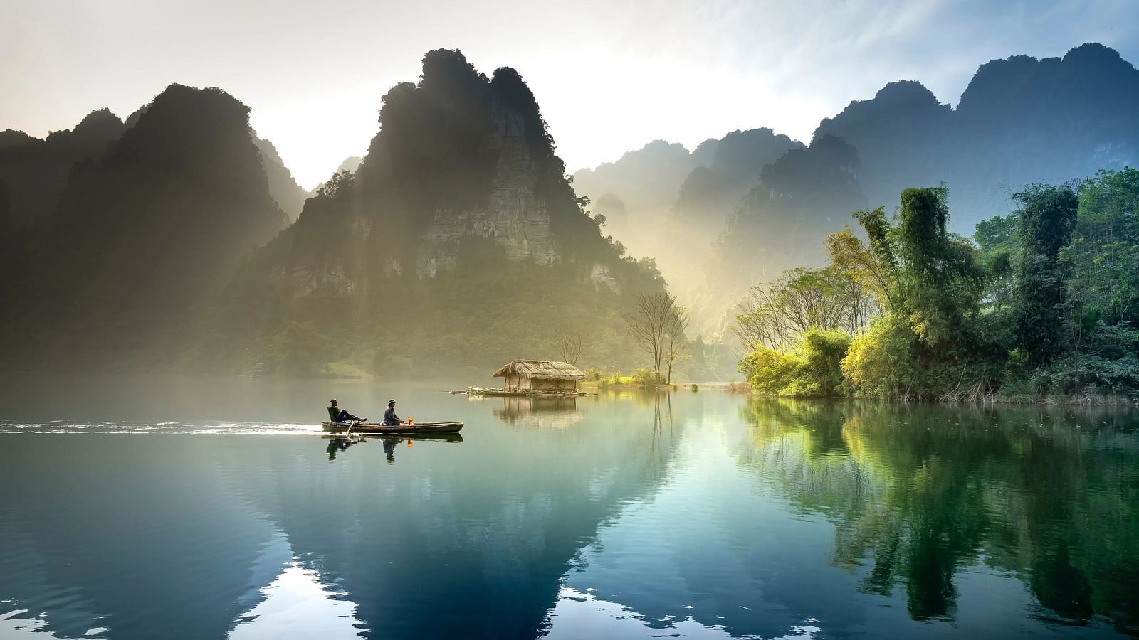 Silhouetted figures in a boat on a reflective lake with hills at sunrise.