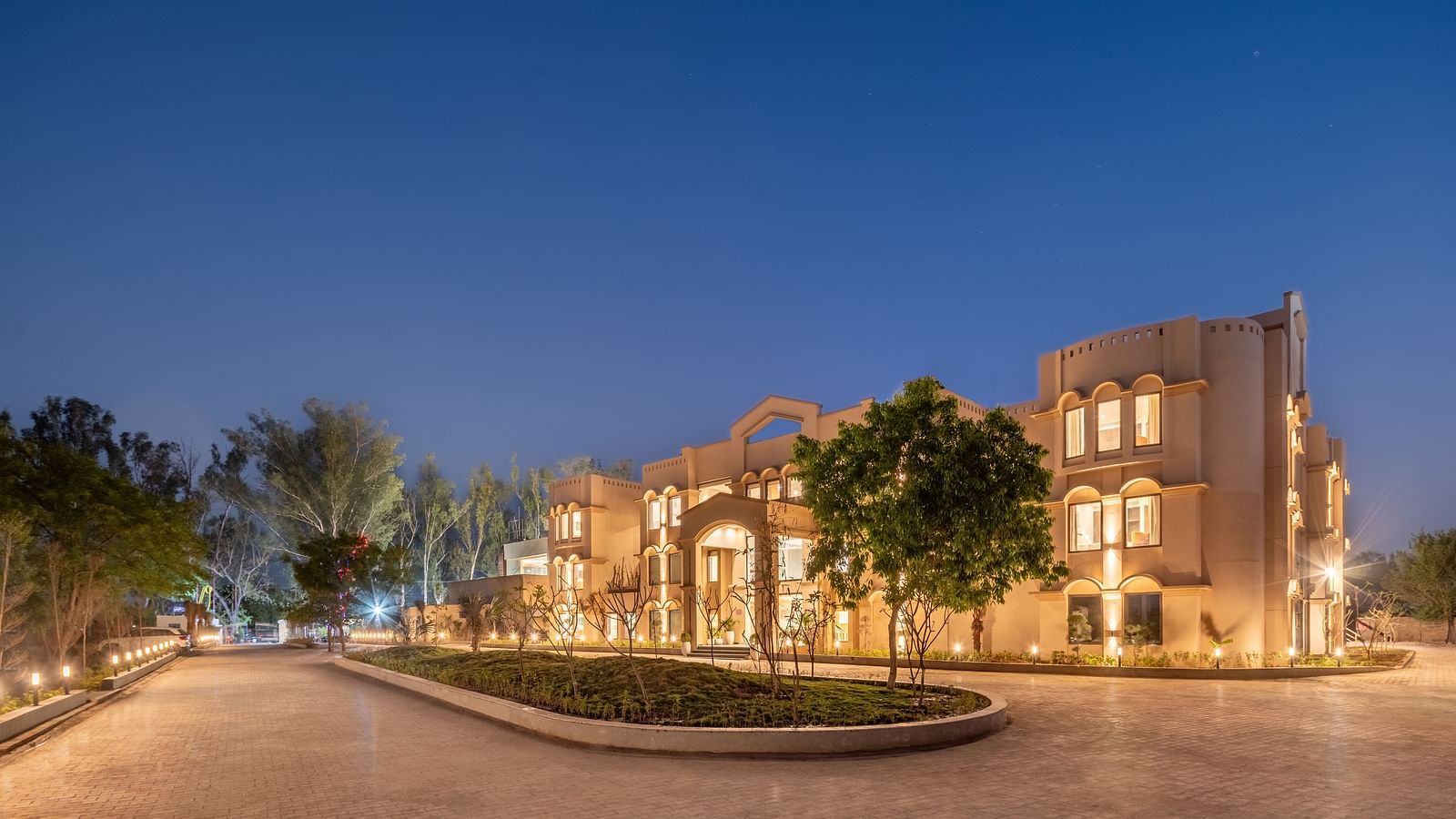 An image of a well lit facade with a huge concrete lawn and a few trees - Brij Eternity, Vrindavan