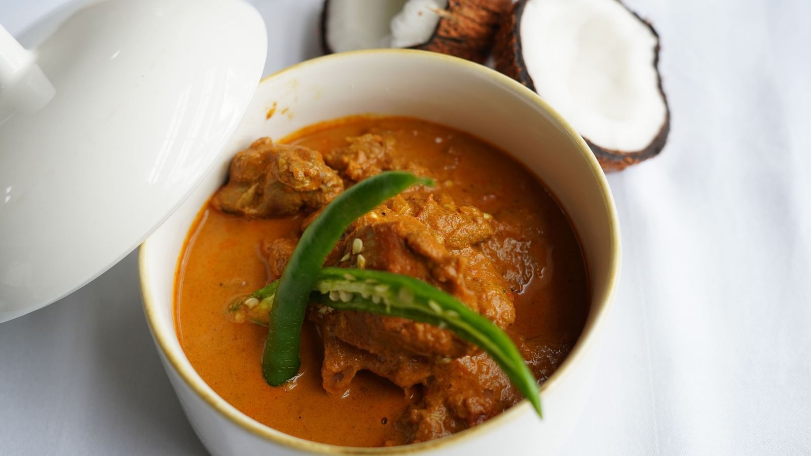 An overview of Chicken Xacuti served in a white claypot with coconut kept next to it.