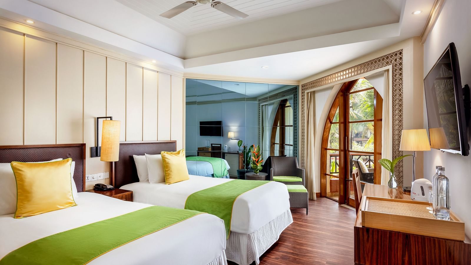 twin beds opposite a wall mounted tv with a ceiling fan in view inside one of the rooms - Caravela Beach Resort Goa