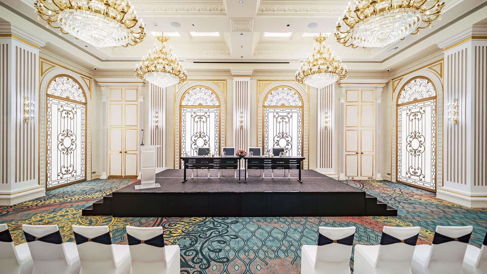 An elegant event space featuring a raised stage area with seating, flanked by rows of white chairs, ideal for presentations or ceremonies, all within a luxuriously appointed hall at Grand Chennai by GRT Hotels.