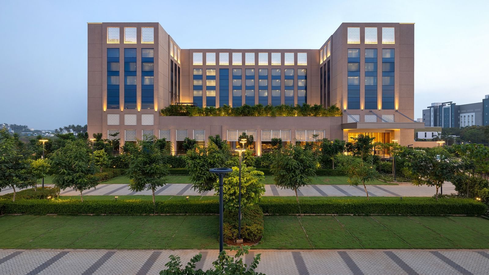 A facade image of our large modern hotel building with numerous windows and manicured landscaping, likely at dusk - Grand Madurai by GRT Hotels