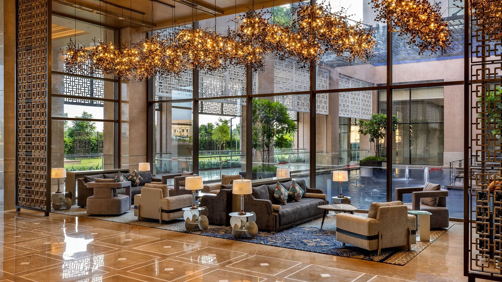A hotel lounge with elegant furniture and a view of outdoor greenery through large glass windows - Grand Madurai by GRT Hotels