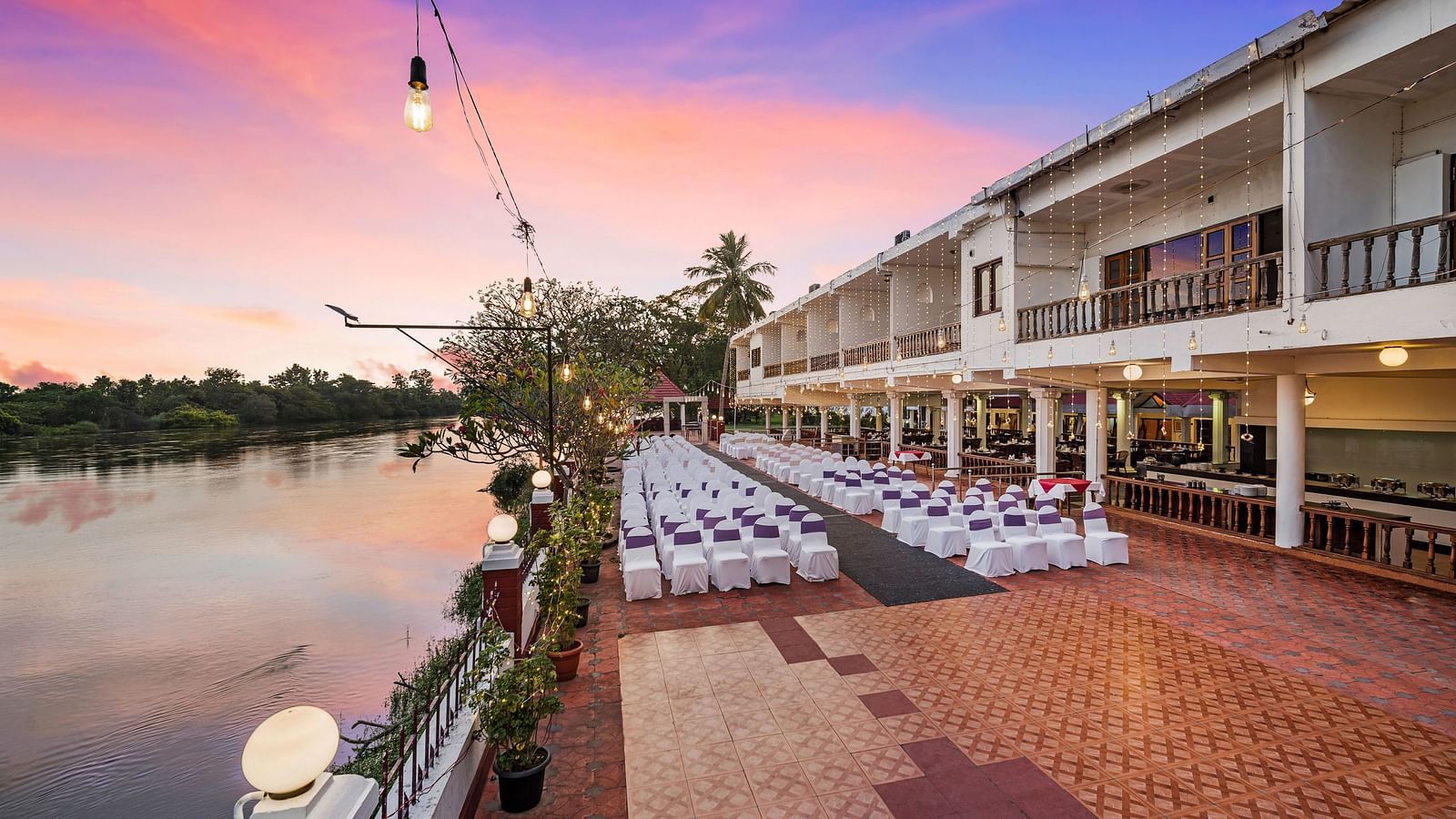 An image of the outdoor event space with seating arrangement near a lake with bright decorative lighting -  GReaT Trails River View Resort Thanjavur By GRT Hotels