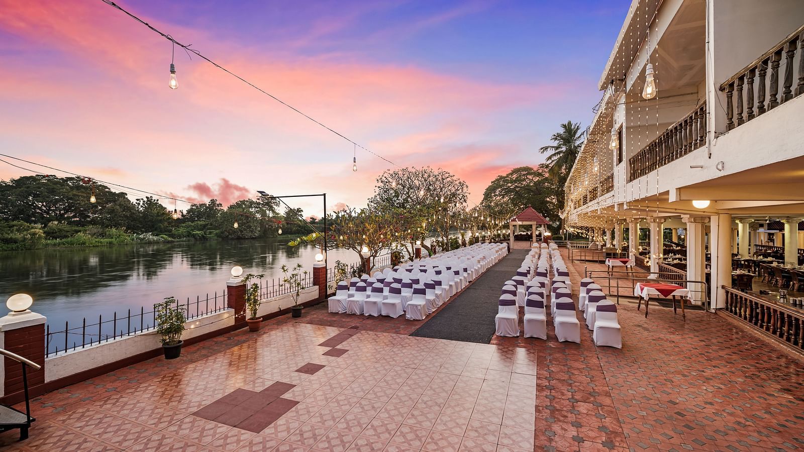 An image of the outdoor event space with seating arrangement near a lake with bright decorative lighting - GReaT Trails River View Resort Thanjavur By GRT Hotels