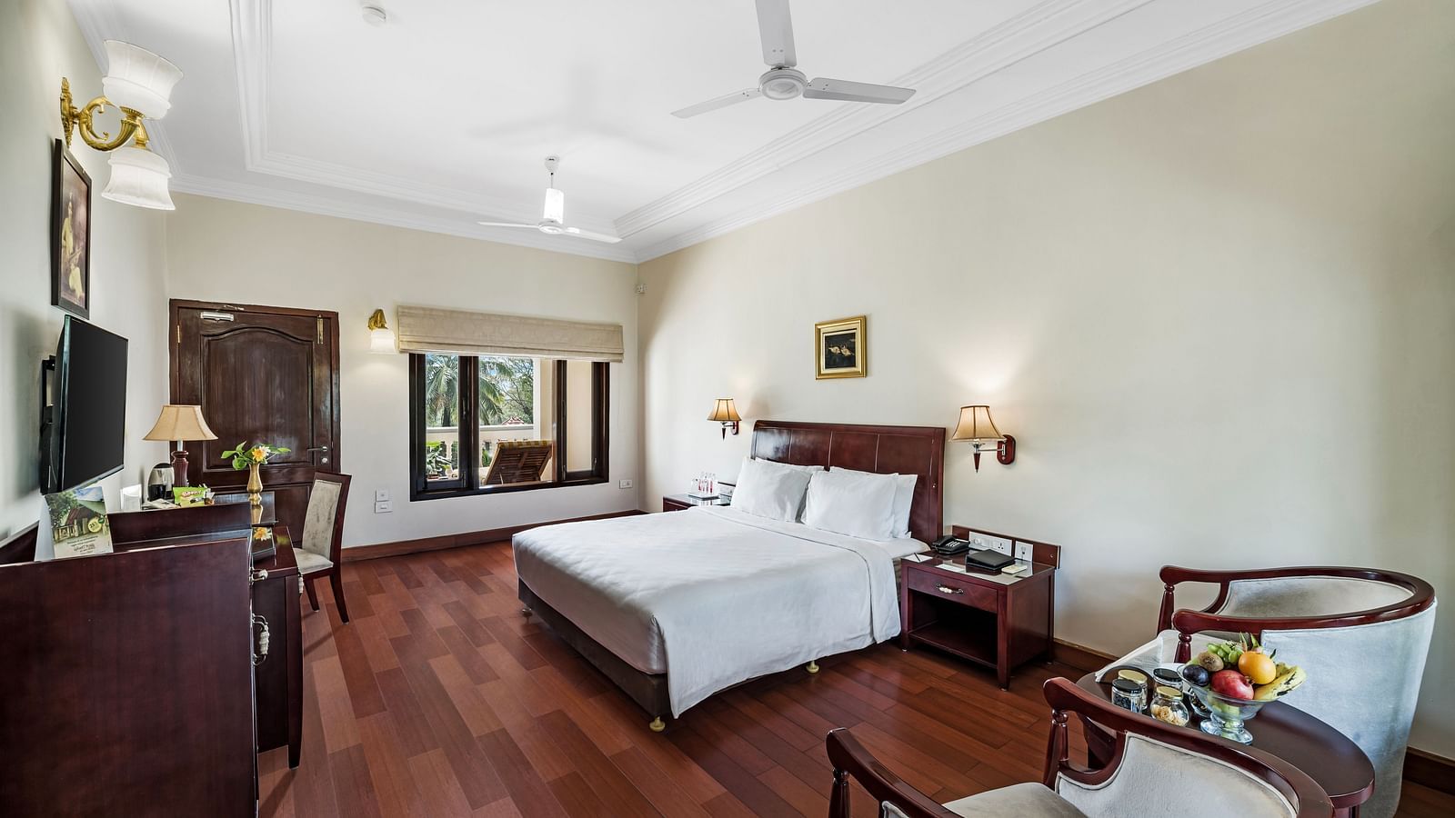 An image of the Garden room with river view featuring a queen size bed, bed side tables, lamps, ceiling fan, flat screen TV, and chairs - GReaT Trails River View Resort Thanjavur By GRT Hotels