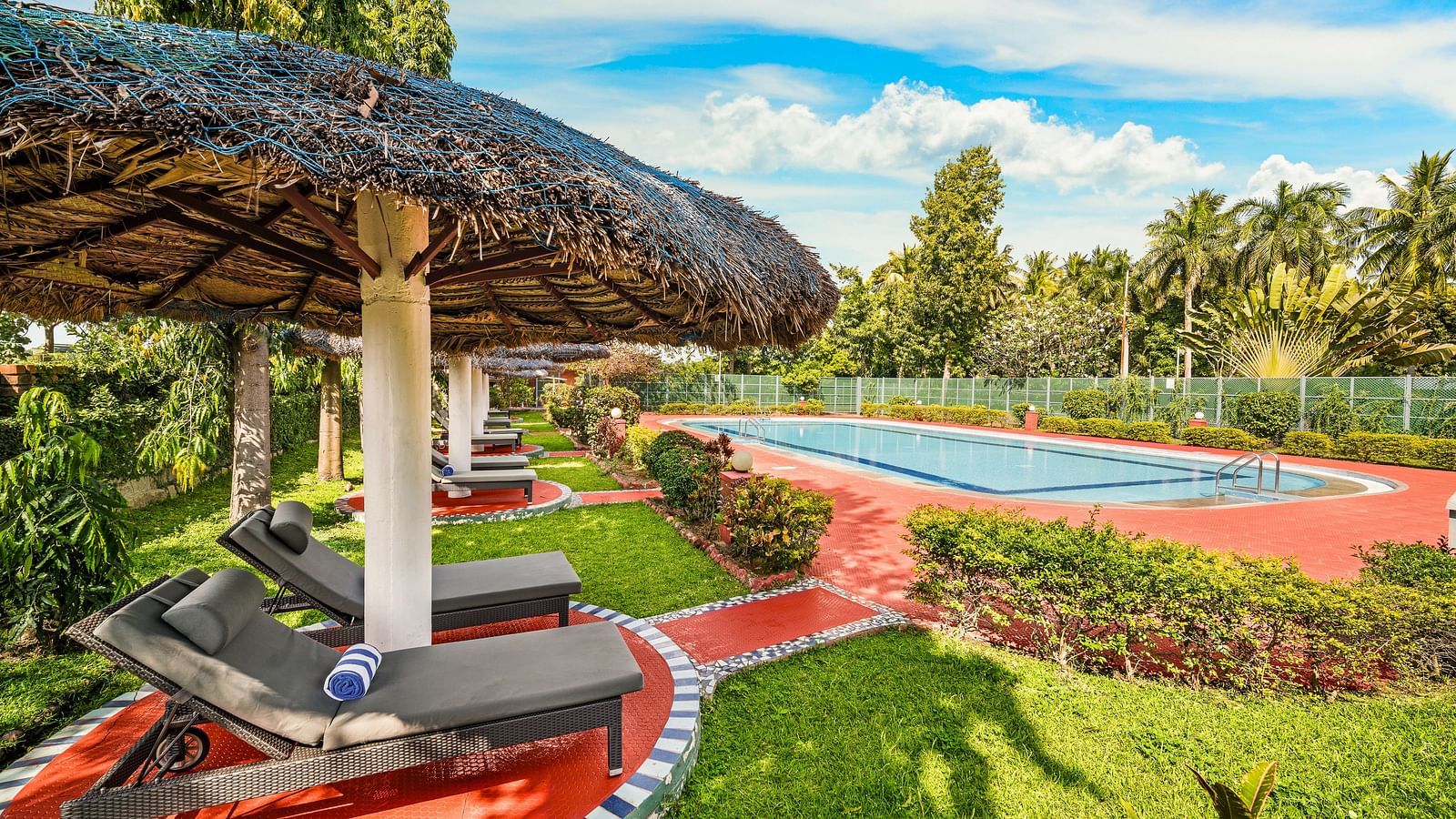 An image of the Kulam Swimming Pool with lounge chairs with thatched roof surrounded by lush greenery - GReaT Trails River View Resort Thanjavur By GRT Hotels