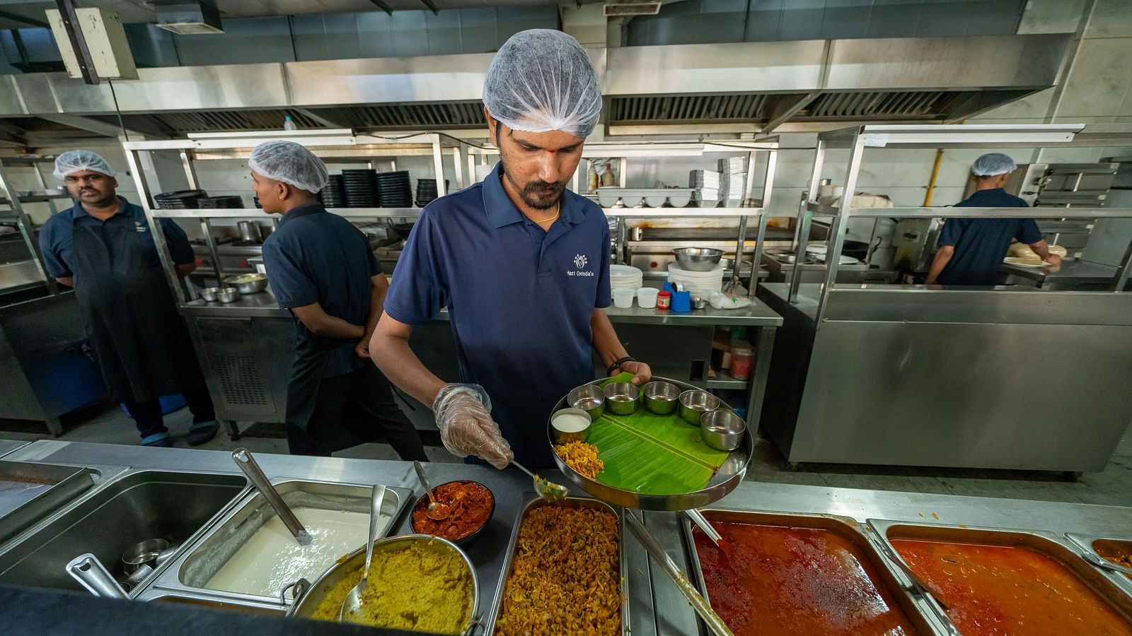 A person serving food from the counter area with one hand while holding a plate in the other at Hari Govinda's