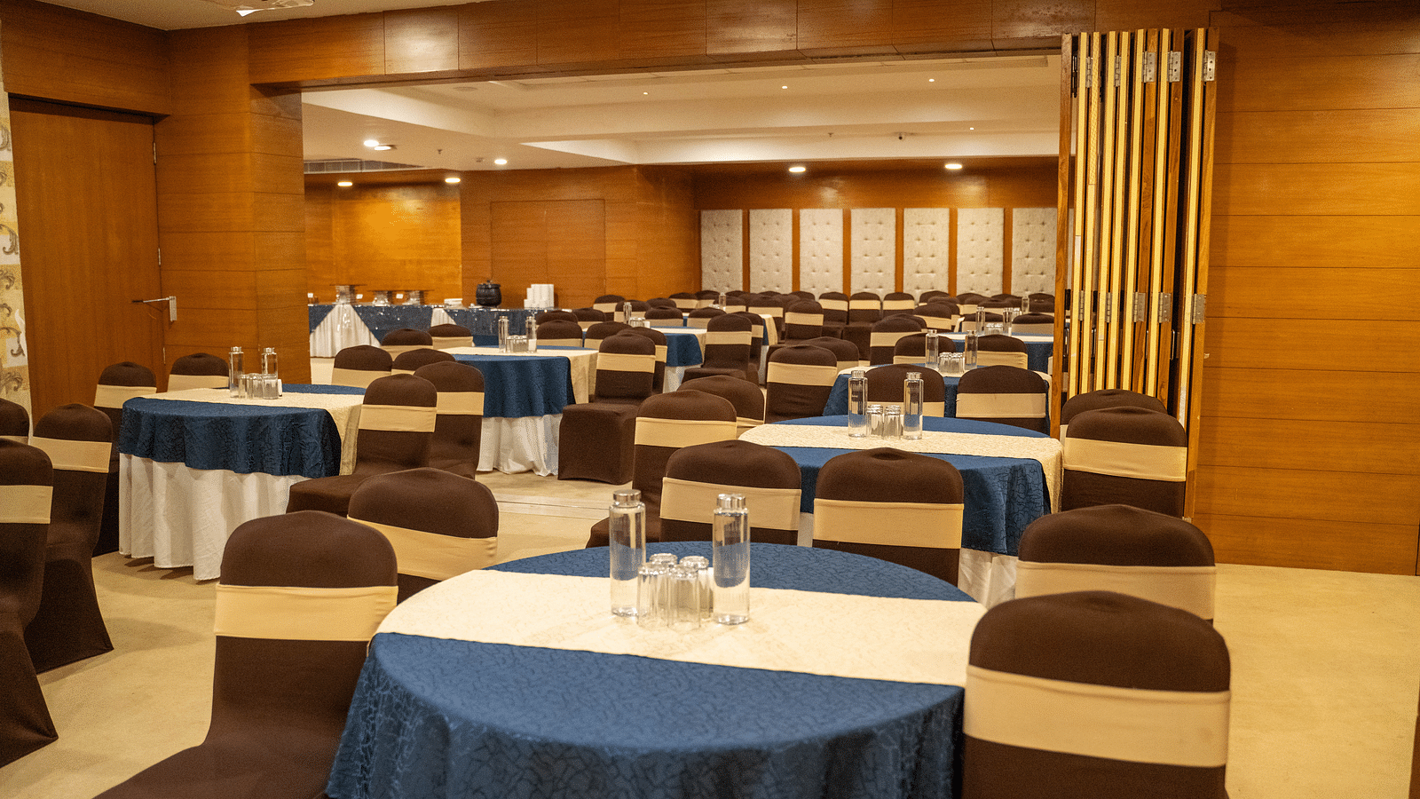 A banquet hall with round tables draped in blue tablecloths and brown chairs, arranged for an event - Hotel Udai Median, Udaipur.