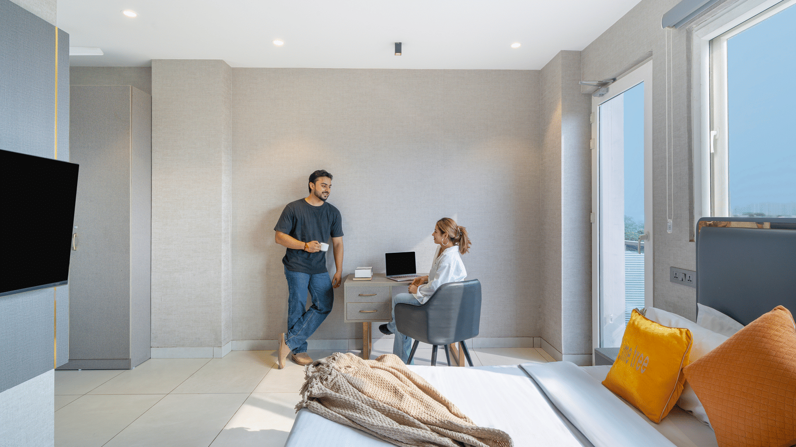 Family with luggage in a bedroom of Lime Tree Hotels & Service Apartment Private Limited featuring a wall mounted television and orange cushions.