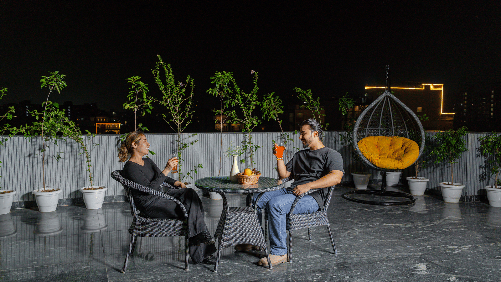 People seated at a rooftop lounge area of Lime Tree Hotels & Service Apartment Private Limited featuring string lighting and a hanging chair at night.