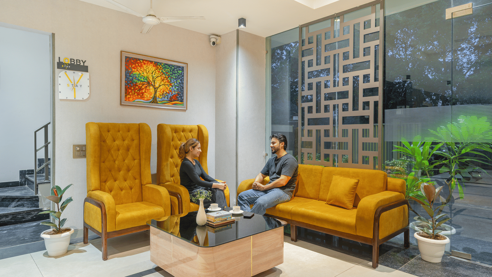 People seated on a yellow sofa in a lounge area of Lime Tree Hotels & Service Apartment Private Limited featuring wall art and potted plants.