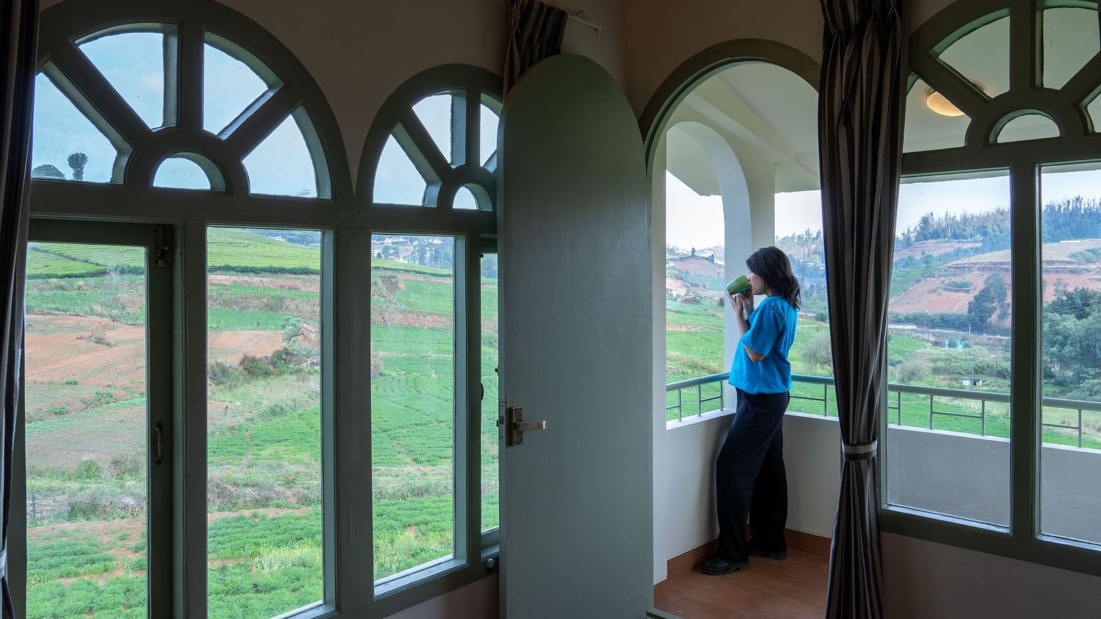 A woman sipping on coffee and looking at the greenery from inside bedroom villa with a balcony | Mango Hill Shola Ooty