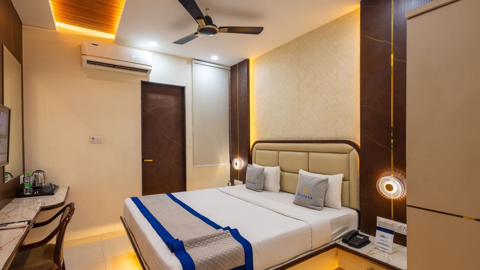 A room with a double bed, warm lighting, blue & beige runner, bedside lamps & a chair facing the wall at Pravasa Stay Dewas Naka, Indore.