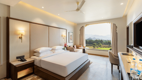 A well-lit bedroom featuring a bed with white linens, a ceiling fan, and a large window looking out onto the greenery at EsthereaRaj Leela, Ranakpur.