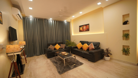 Living room of Lime Tree Hotels & Service Apartment Private Limited featuring a black sofa, a wall mounted television, and dark curtains.