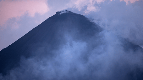 The towering dark peak of Barren Island volcano shrouded in swirling smoke and twilight clouds.