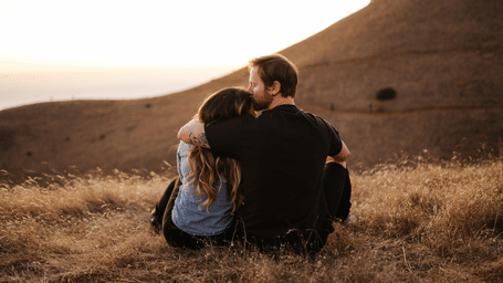 A couple sitting with their arms around each other on a dry, grassy hillside, watching a hazy sunset.