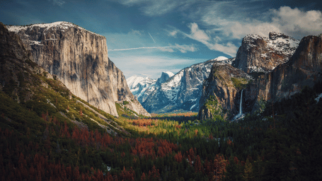 A dramatic view of Yosemite Valley with towering granite cliffs, including El Capitan, snow on the peaks, and autumn colors in the forest.