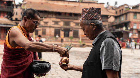 A local devotee offering alms into a Buddhist monk's bowl at a historic square temple complex in Kathmandu Valley, Nepal.