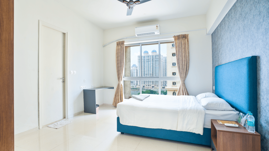 A bright hotel bedroom with a blue headboard, a white double bed, and a large window overlooking urban tower blocks at Apartel by Aarin, Oragadam.