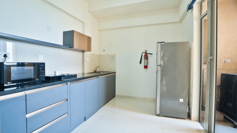 A modern kitchen with blue cabinetry, a grey fridge, and a microwave, adjacent to a bright glass door leading outside at Apartel by Aarin, Oragadam.