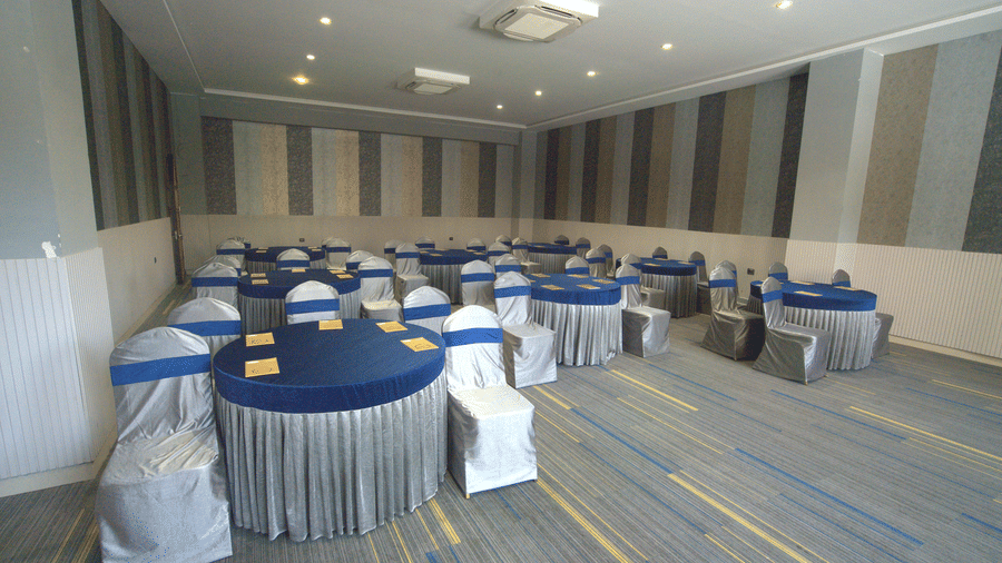 An angled shot of an elegant function room featuring several circular tables with blue linens and chairs covered in silver fabric at Apartel by Aarin, Oragadam.