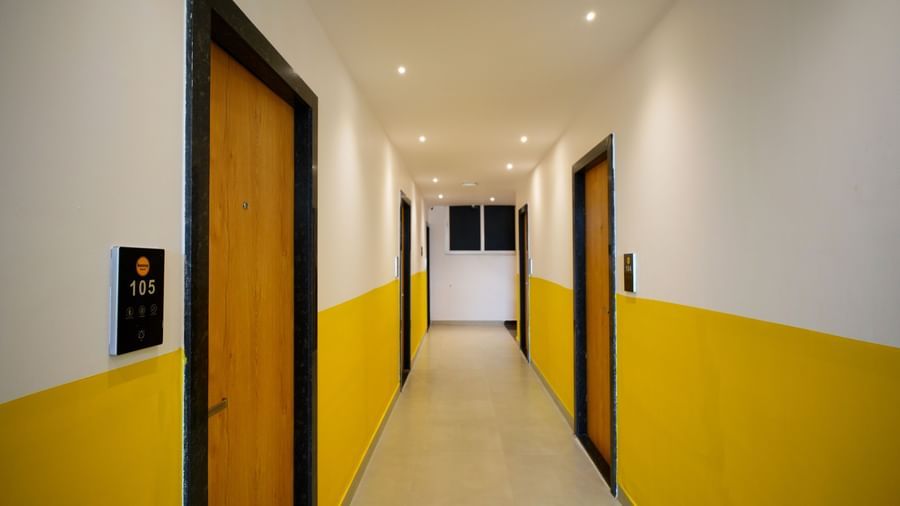 A hallway with yellow and white walls, featuring brown doors and recessed lighting, leading towards a bright area in the distance  - Beestay Hotels Financial District, Hyderabad 