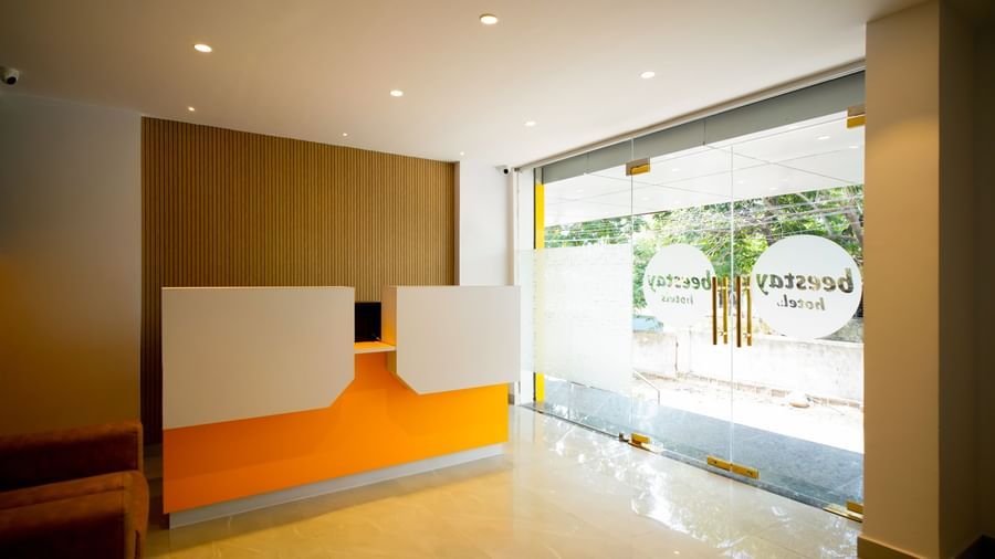Side view of a modern hotel lobby with a bright orange and white reception desk, 2 brown armchairs, and a warm-toned wall behind them - Beestay Hotels Financial District, Hyderabad 