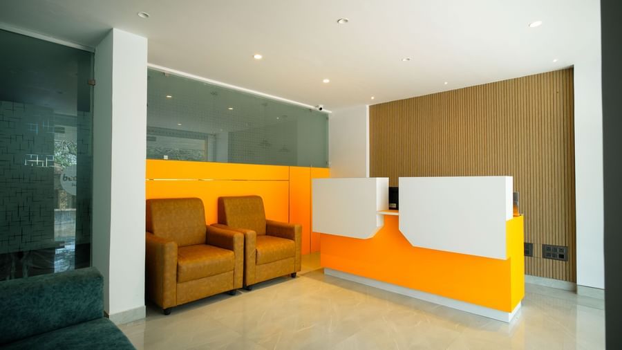 A modern hotel lobby with a bright orange and white reception desk, 2 brown armchairs, and a warm-toned wall behind them - Beestay Hotels Financial District, Hyderabad 