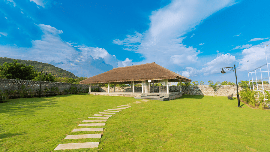 An expansive green lawn with a tile pathway leading towards a stage area with thatched roof inside Darza Resorts, the best wedding resort in Coimbatore.