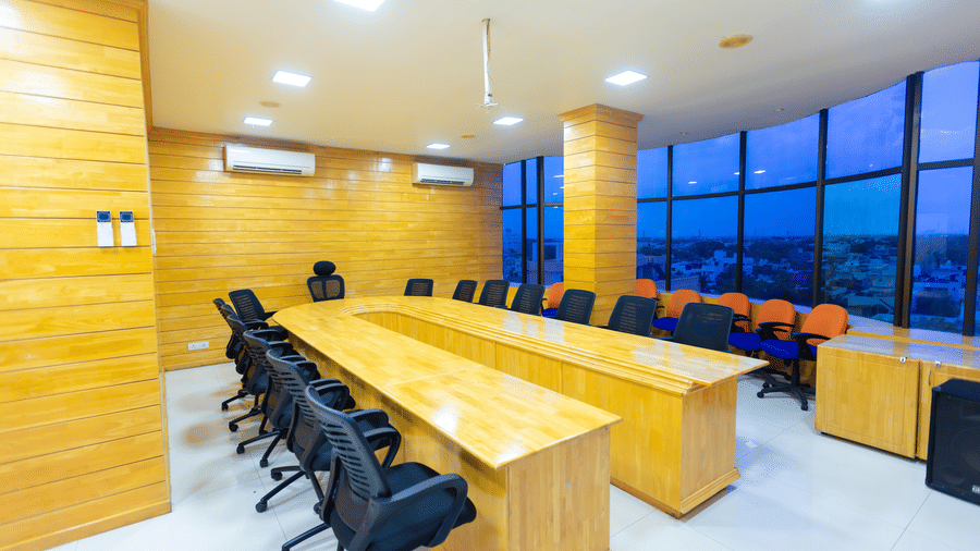 The business centre at DSF Grand Plaza, Tuticorin with a U-shaped conference table, black, orange, and blue chairs, and a curved window wall overlooking the city at dusk.
