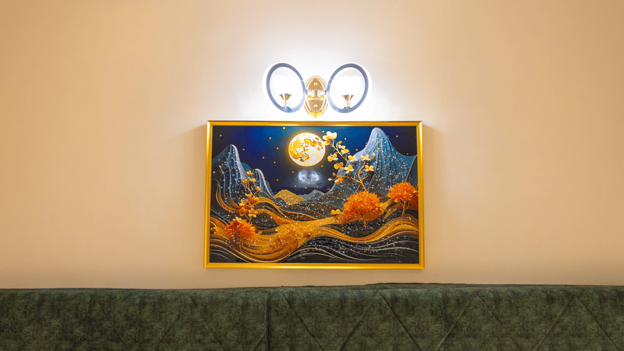 Decorative wall art at DSF Grand Plaza, Tuticorin above a green quilted headboard, showing a moonlit mountain scene with a double-ring wall lamp overhead.