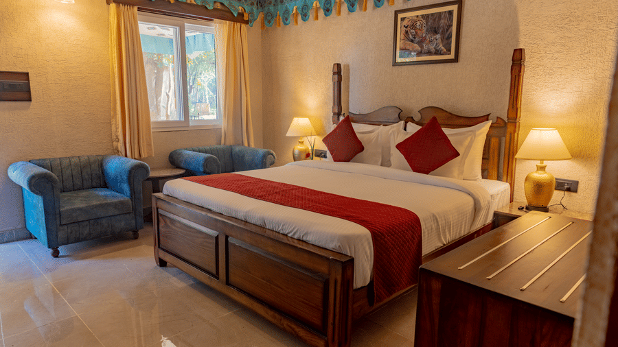 Guest bedroom at Estherea Bagh, Ranthambore, showing a wooden bed with accented bedding, upholstered armchairs, and drawn curtains.